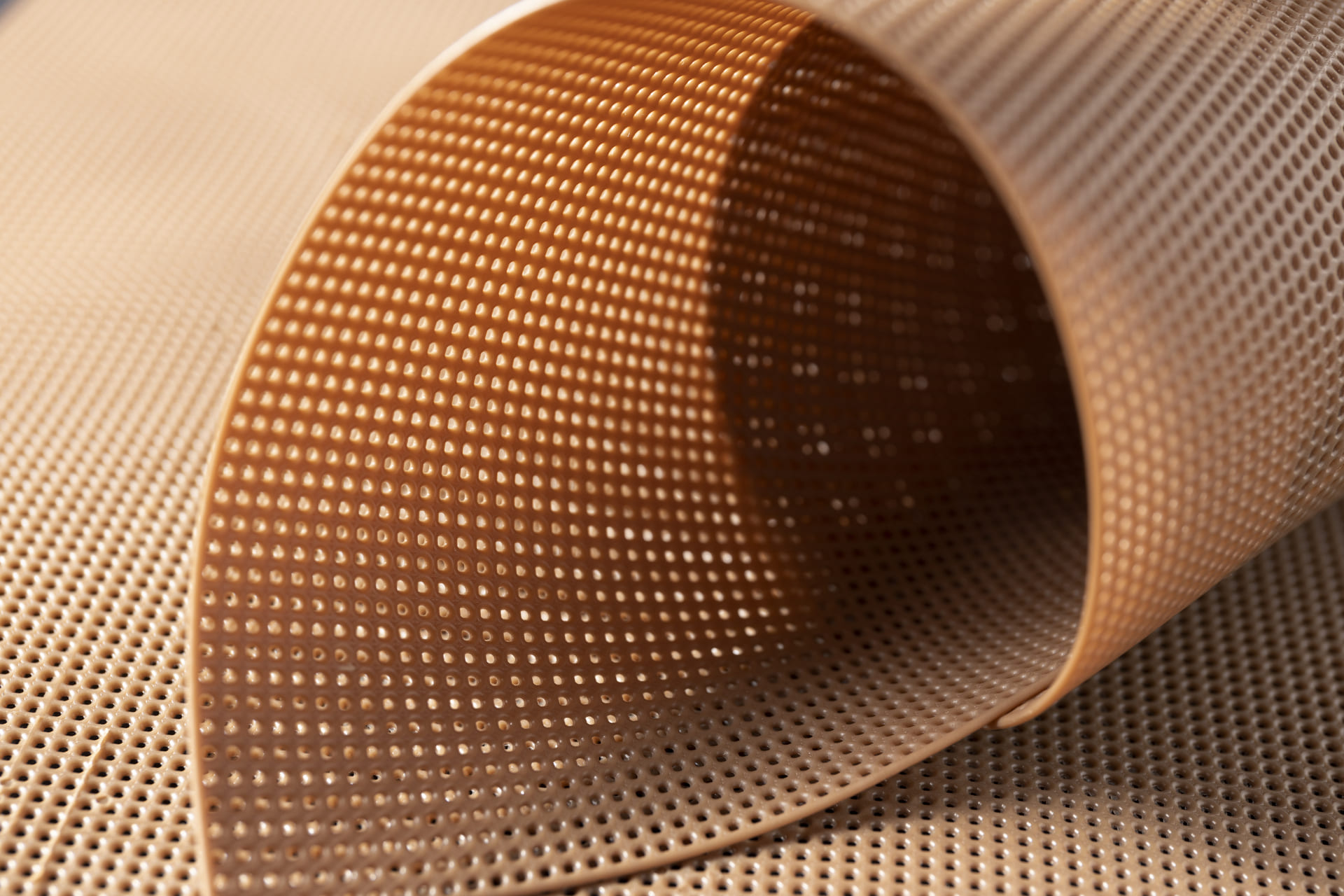 Close-up de um material de malha marrom flexível e perfurado moldado em uma curva suave. A textura é destacada pela iluminação, enfatizando os pequenos furos uniformemente espaçados por toda a superfície. Ideal para isolamento acústico, seu fundo exibe uma textura semelhante.